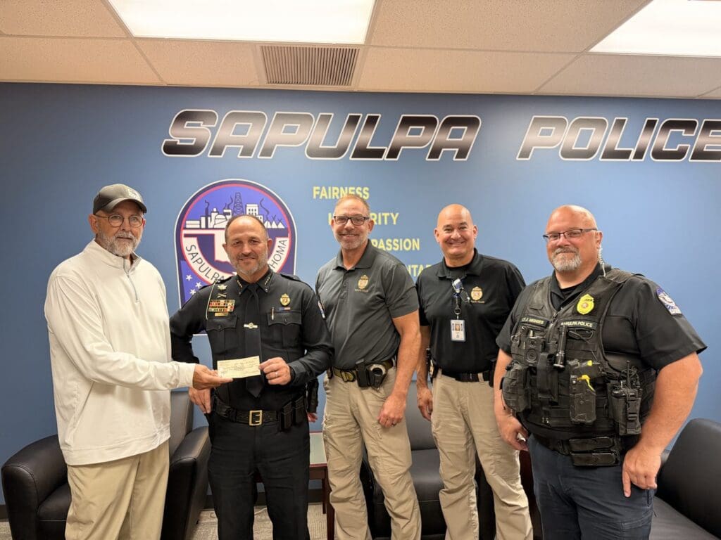 Phil Kennedy hands a check to Sapulpa Police Chief Mike Reed as Deputy Chief Rob Glenn, Major Terry Wiggs and Lt. Jason Hedges look on in front of a Sapulpa Police wall.